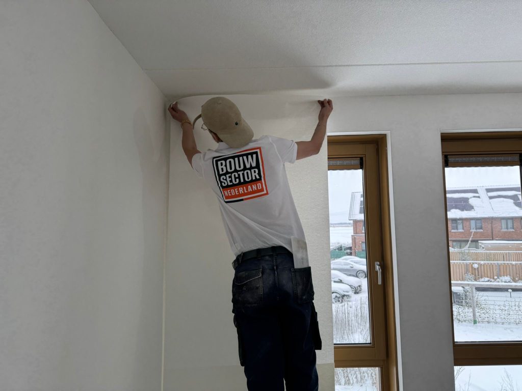 Behanger van Bouwsector Nederland zet een baan vlies strak tegen het plafond in een nieuwbouwkamer met houten kozijnen