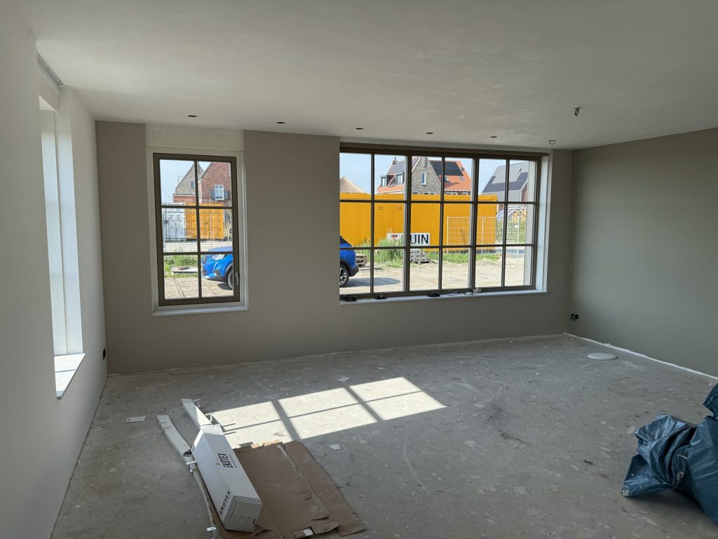 Nieuwbouwkamer met wanden in een warme grijstint en een groot roedenraam dat uitkijkt op nieuwe huizen