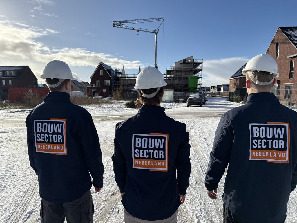 Drie medewerkers van Bouwsector Nederland met witte helm in een besneeuwde straat met nieuwbouw en een bouwkraan